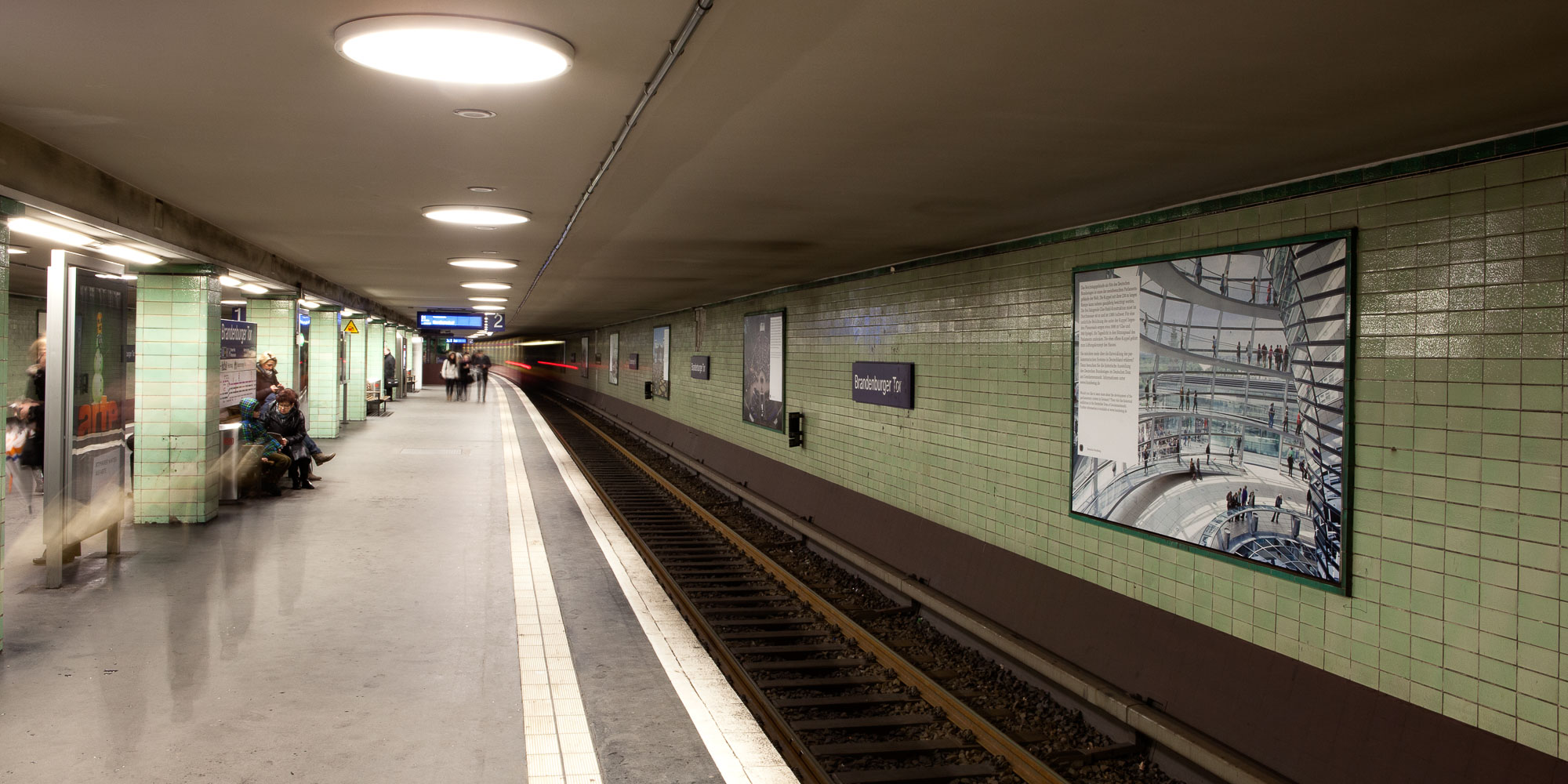 Blick auf den Bahnsteig, an der Wand rechts sind verschiedene Plakate angebracht. Mehrere Personen warten auf dem Bahnsteig, von weit hinten fährt eine S-Bahn ein.
