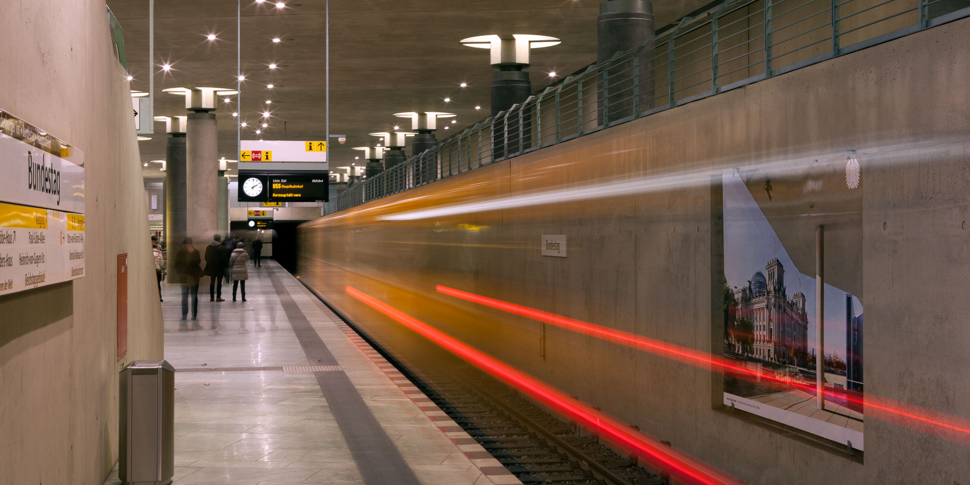 Blick auf den Bahnsteig, an der Wand rechts ist ein Plakatbeispiel zu sehen. Mehrere Personen warten auf dem Bahnsteig, eine U-Bahn fährt mit Geschwindigkeit aus dem Bahnhof.